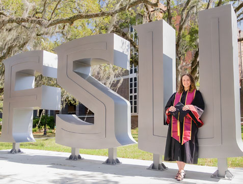 Picture of student wearing gown holding her cap at statue of FSU letters.